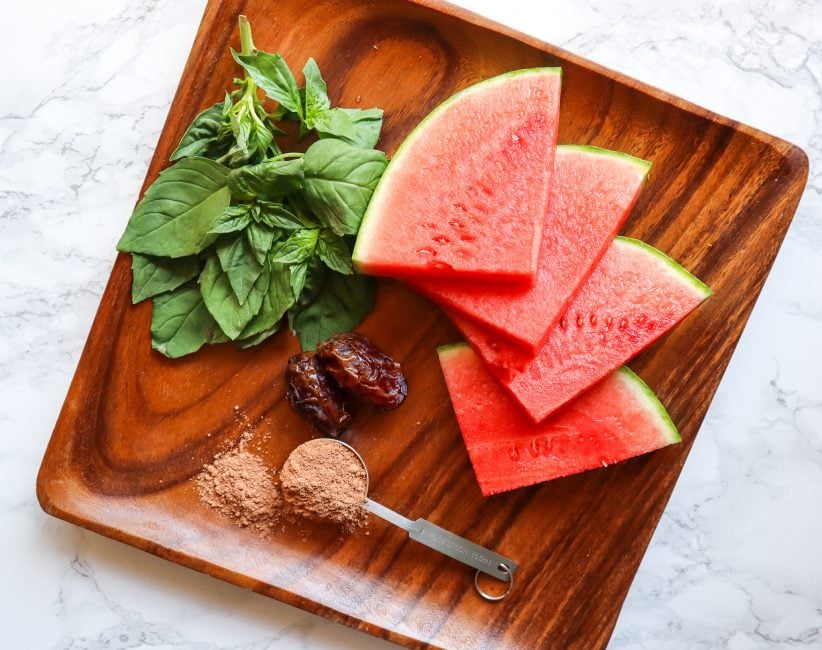 ingredients for watermelon smoothie on a wooden plate shown are sliced watermelon two medjool date a tablespoon of cacao and fresh basil