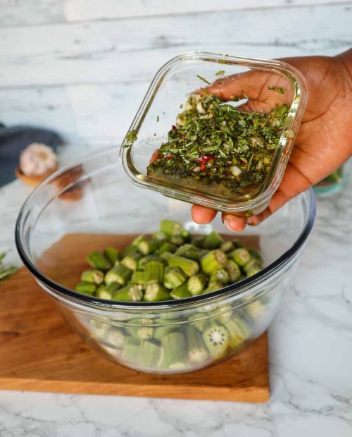 A clear bowl of sliced okra with a hand holding herbs and olive oil over the bowl, ready to pour.